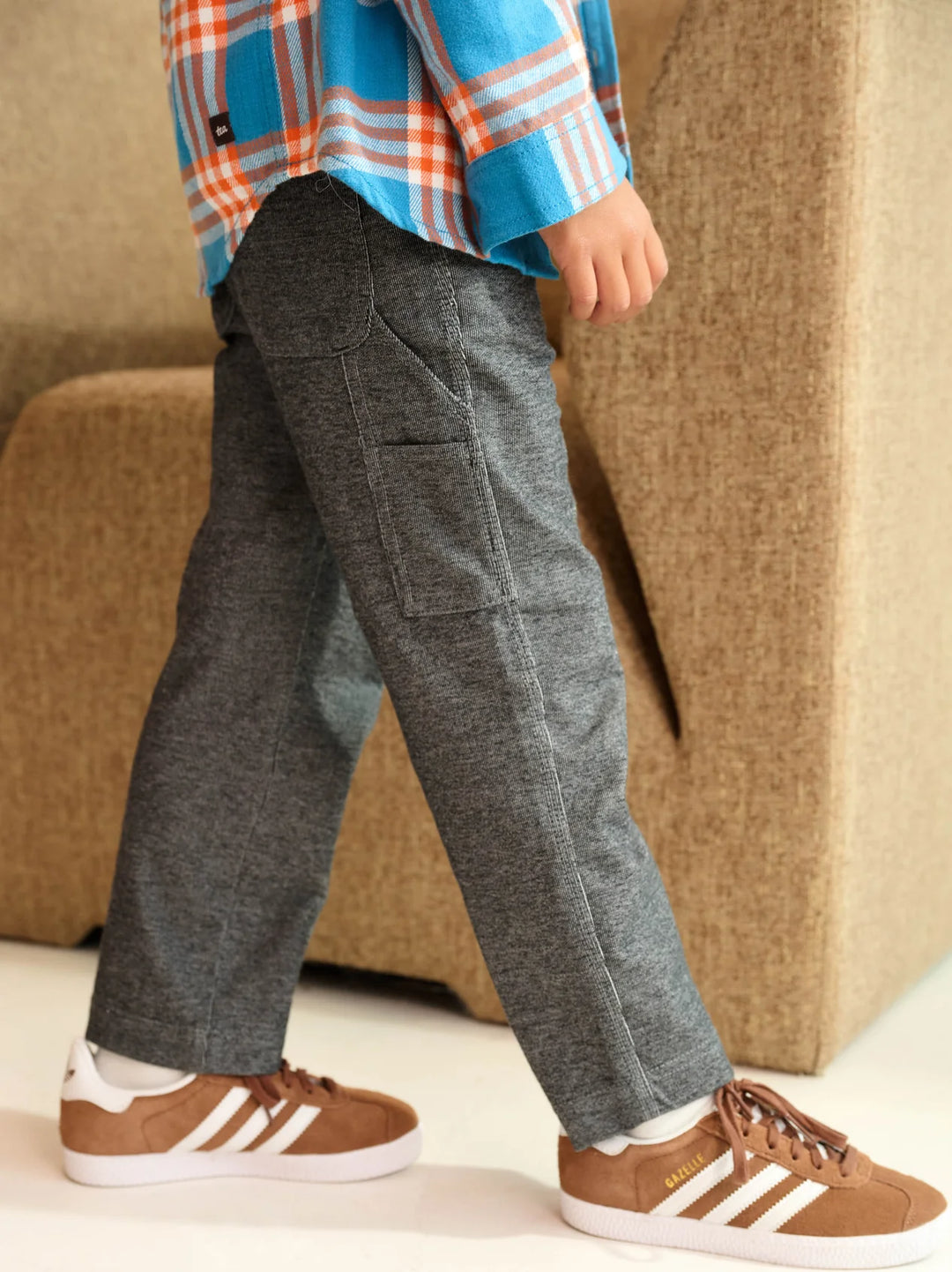 A child wearing dark gray heathered cargo pants, a blue and orange plaid shirt, and brown Adidas sneakers, standing in front of a beige upholstered chair.