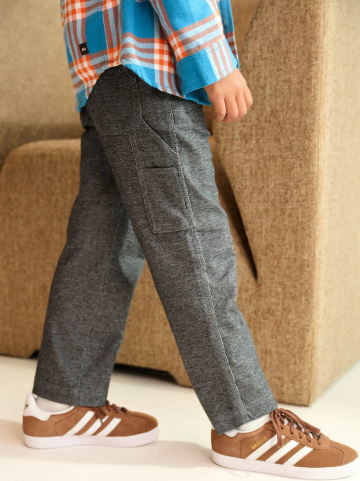 A child wearing dark gray heathered cargo pants, a blue and orange plaid shirt, and brown Adidas sneakers, standing in front of a beige upholstered chair.