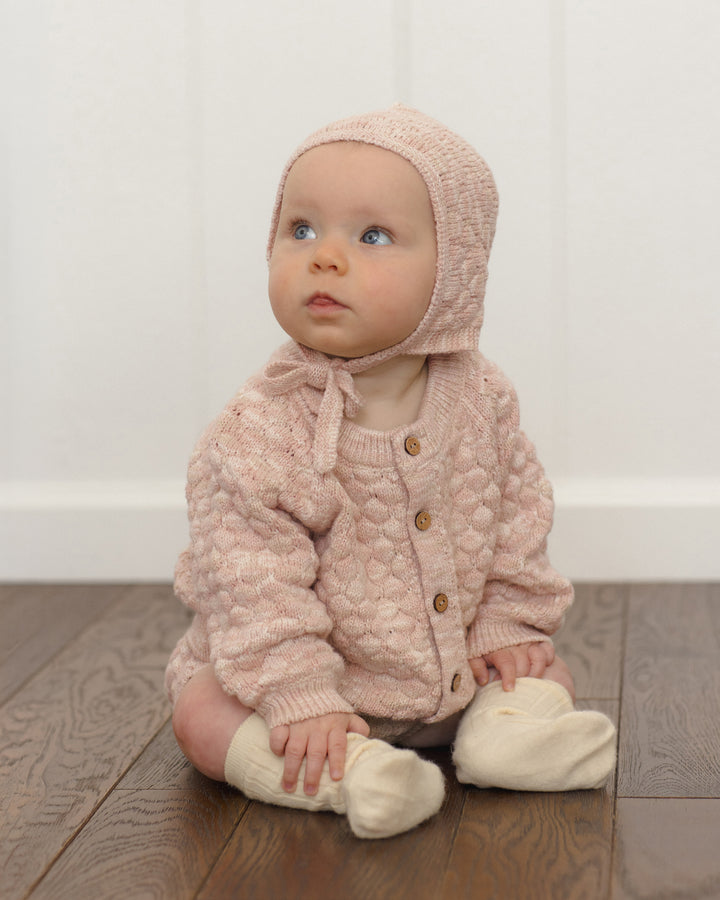 A baby sitting on a wooden floor wearing a light pink textured knit bonnet and matching cardigan with cream booties.