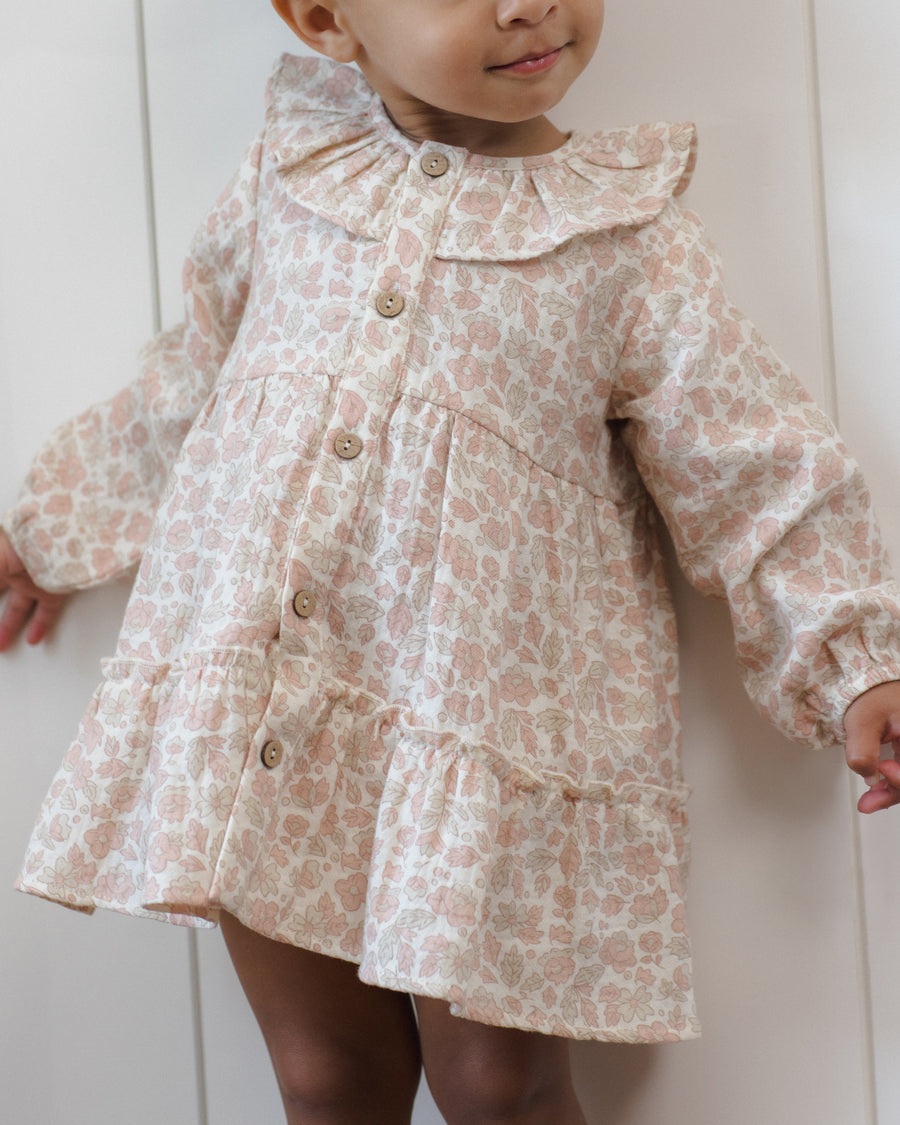 A toddler wearing a long-sleeve ruffled dress with a light pink and beige floral pattern, standing against a white paneled wall.