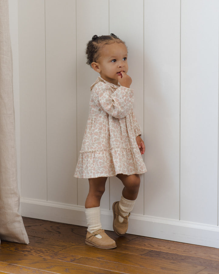 A toddler girl wearing a long-sleeve cream and beige floral print dress, cream socks, and tan mule shoes, standing against a white paneled wall on a wooden floor.