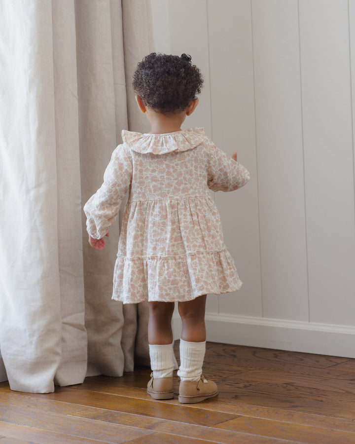 A toddler girl seen from behind wearing a cream and pink floral print long-sleeve dress with a ruffled collar, standing on a wooden floor next to a beige curtain.