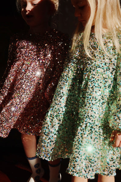 Two little girls standing in sequin dresses. One little girl in gold and green sequins and the other in pink sequins. 