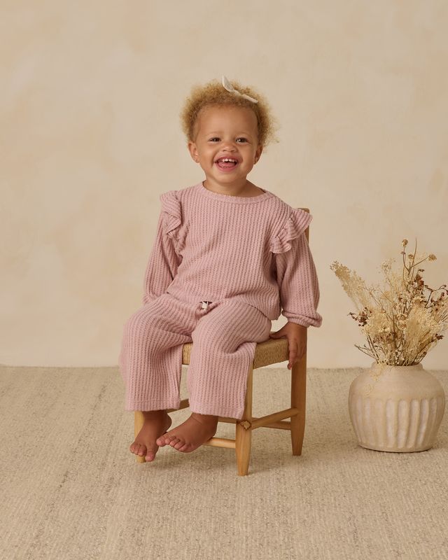 Child wearing a pink ribbed outfit sitting on a wooden stool with a vase of dried flowers in the background.