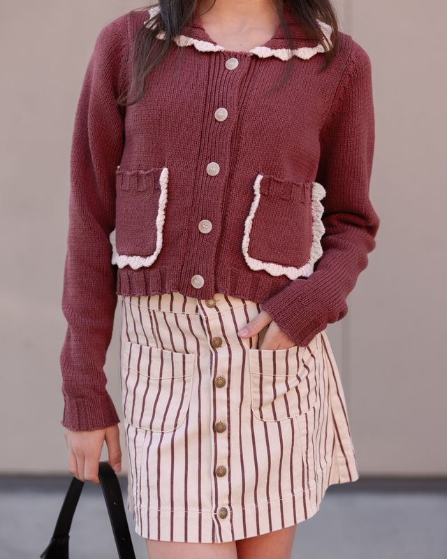 Person wearing a maroon cardigan with lace trim and a striped skirt against a neutral background