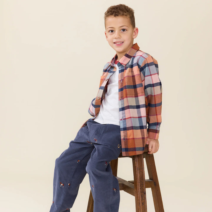 boy sitting on a stool wearing a rust plaid shirt open with a t-shirt open. 