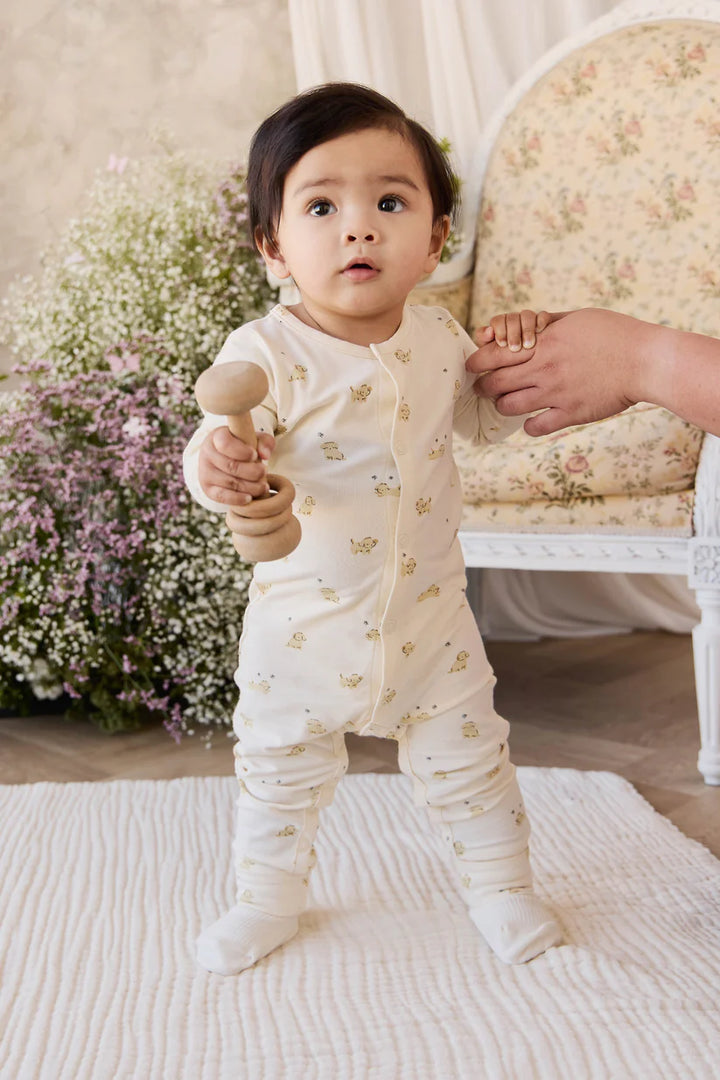 Baby in a white outfit with puppies on it standing on a rug with and a chair in the background