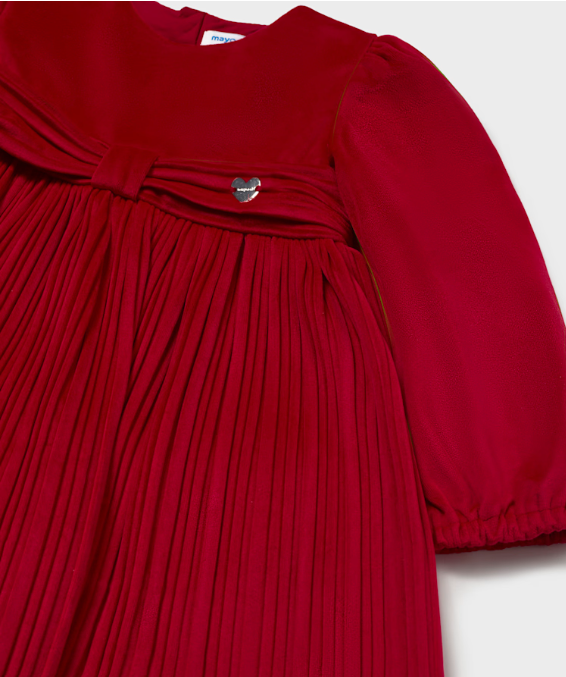 A close-up of a red dress featuring a pleated skirt, a gathered waist with a bow detail, and a small silver heart-shaped brooch, set against a plain light background.