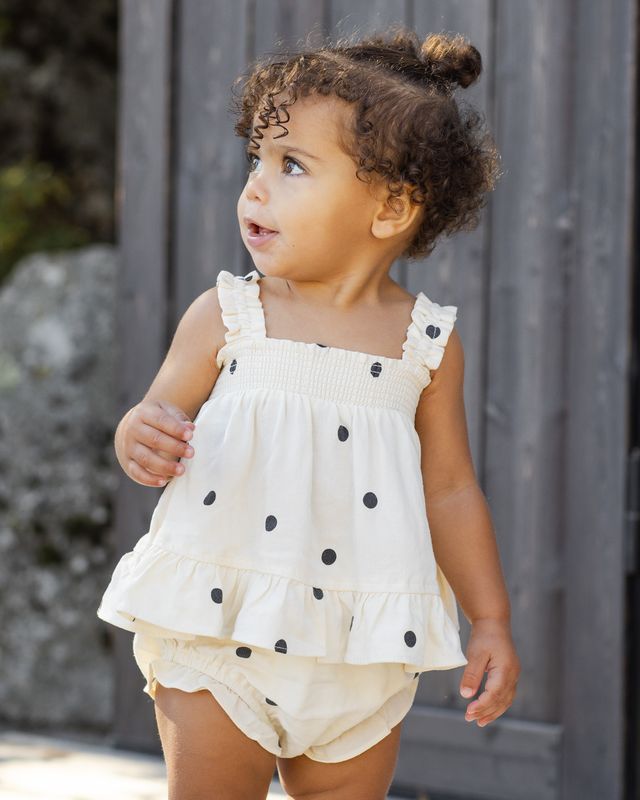 Child wearing a white top with black polka dots and beige shorts outdoors.
