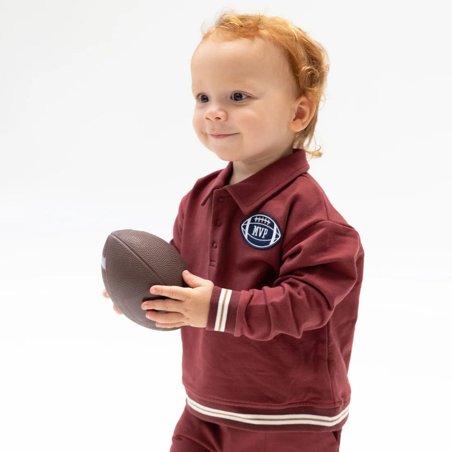 A toddler with curly red hair stands against a white studio background, holding a brown football. The child is wearing a burgundy long-sleeve polo-style top with white stripes on the cuffs and hem, plus a circular chest patch reading 'Team MVP'.
