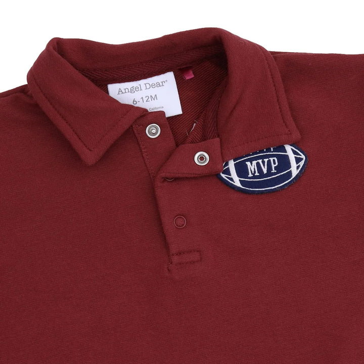 A close-up of a burgundy polo shirt featuring a white neck label that reads 'Angel Dear 6-12M' and a navy blue MVP football patch on the chest.