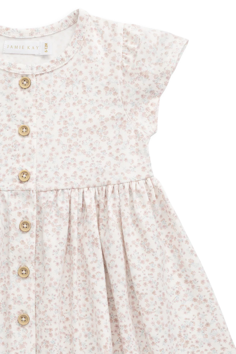 Close-up of a children's dress with a small floral print in soft pink and beige on an off-white base, featuring a button-front bodice and short flutter sleeves.