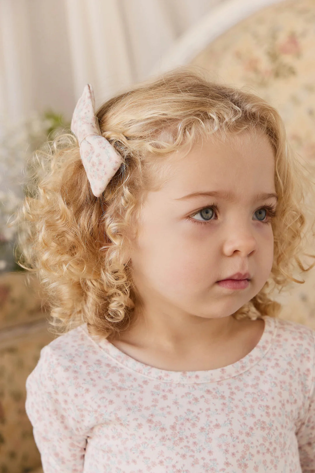 Young girl with curly blonde hair wearing a pink bow, looking off to the side.
