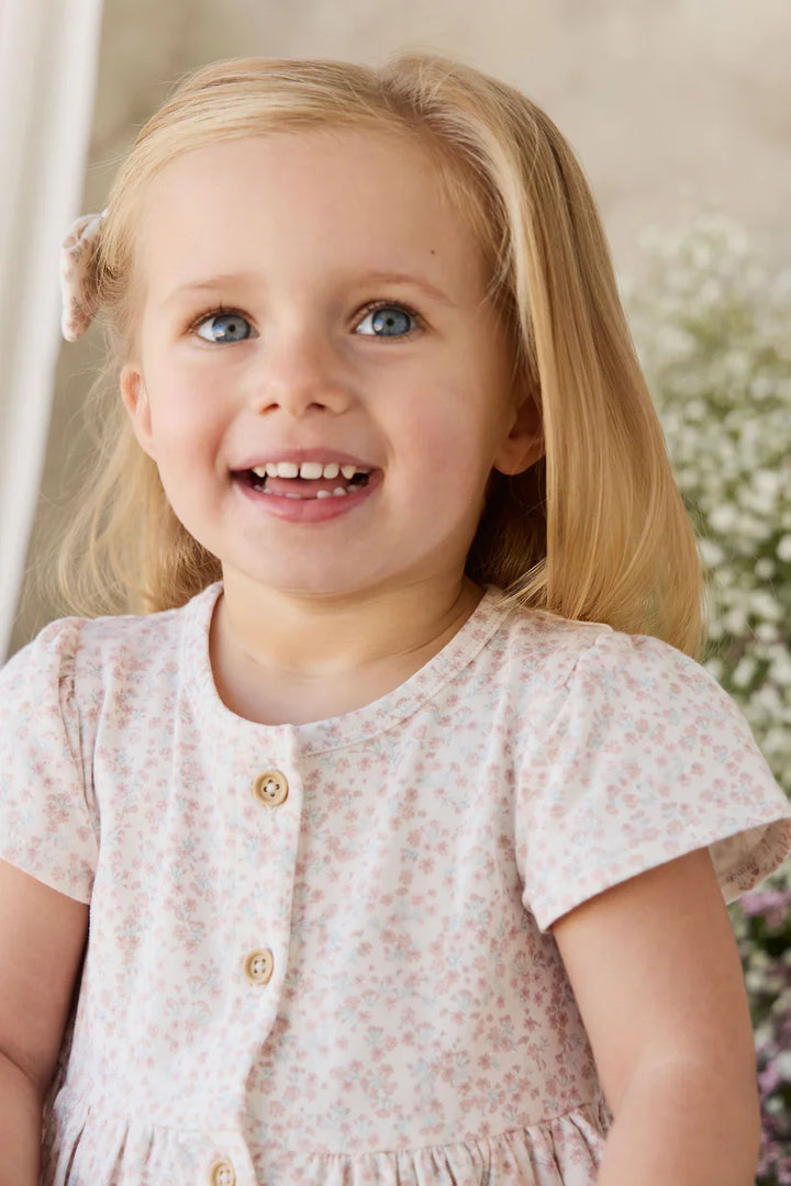 A young blonde girl with blue eyes smiling, wearing a short-sleeve light pink and white floral dress with gold buttons.