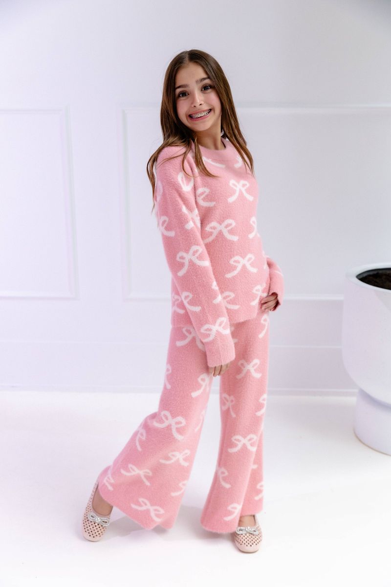 A young girl wearing a pink fuzzy two-piece sweater set with a white bow pattern, standing in a bright white room.