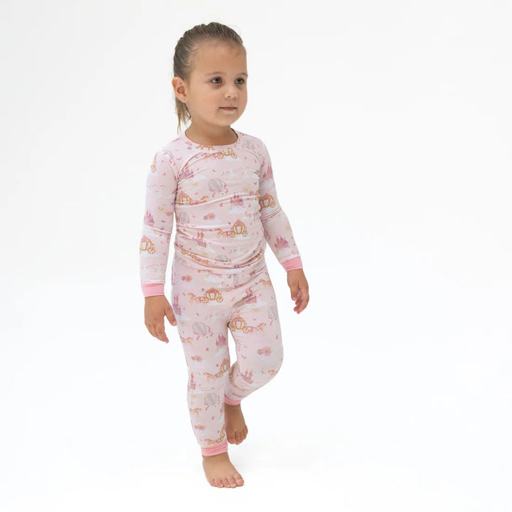 A young child wearing a light pink long-sleeve pajama set with a whimsical castle and fairy print, walking barefoot against a plain white background.