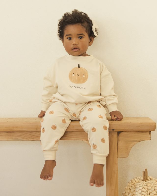 Child wearing a cream-colored outfit with pumpkin design sitting on a wooden surface.