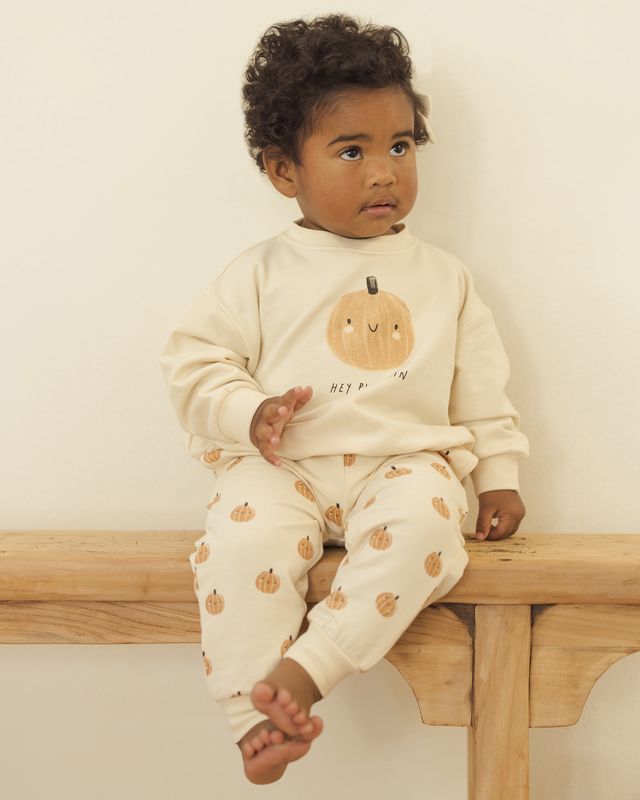 Child wearing a cream-colored outfit with pumpkin patterns, sitting on a wooden bench.