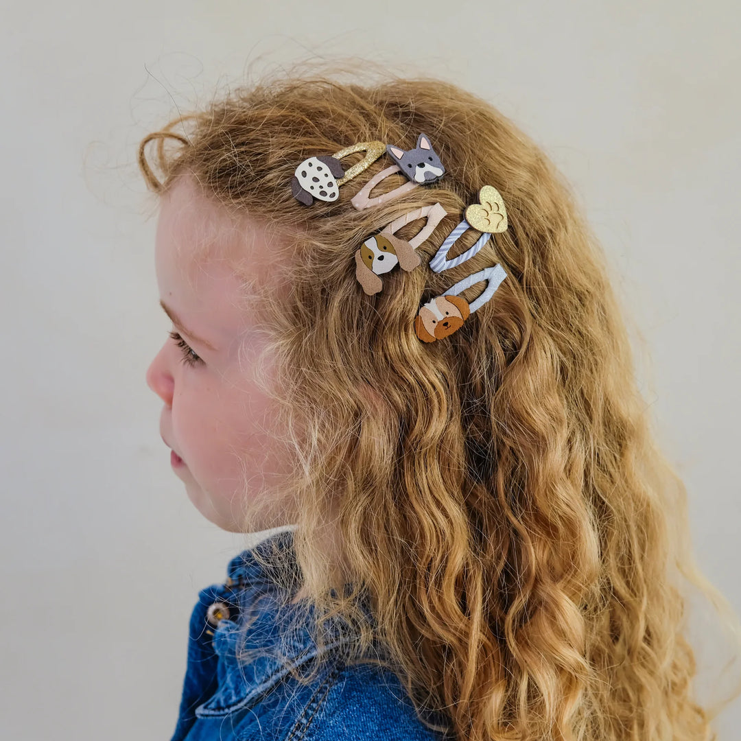 A side-profile view of a young girl with long, wavy blonde hair wearing six small animal-themed hair clips in pastel colors, set against a plain light gray background.