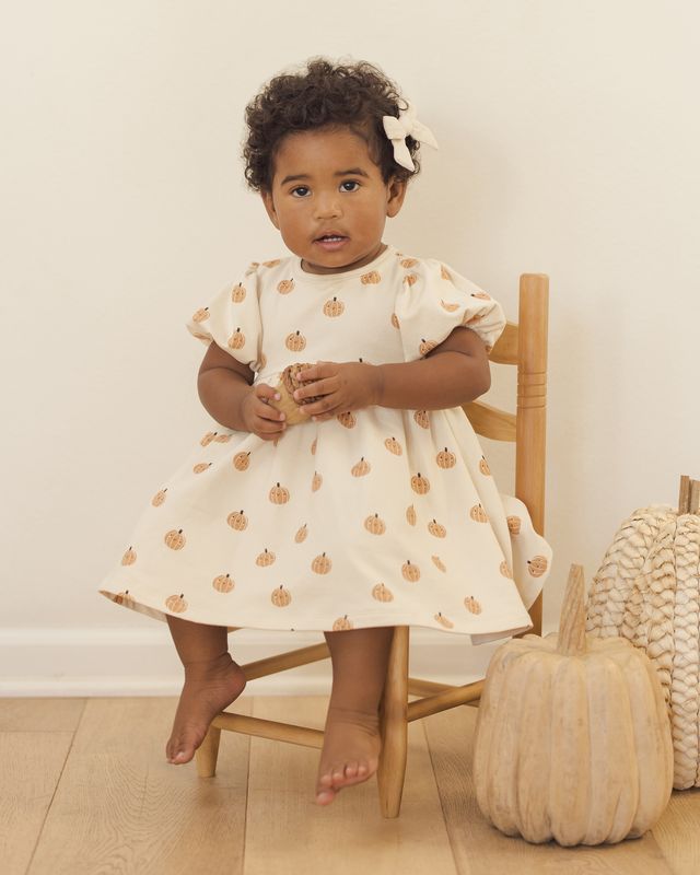 Young child in a beige dress with pumpkin pattern sitting on a chair next to pumpkins.