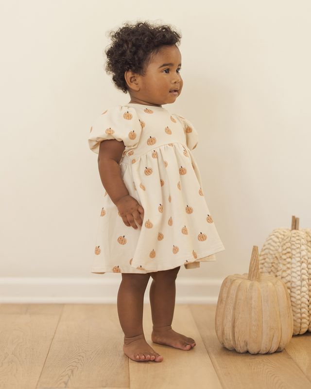 Child wearing a beige dress with pumpkin pattern next to woven pumpkins indoors.