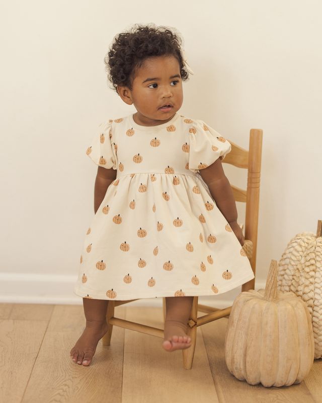Child wearing a beige dress with pumpkin pattern sitting on a chair next to a textured pumpkin.