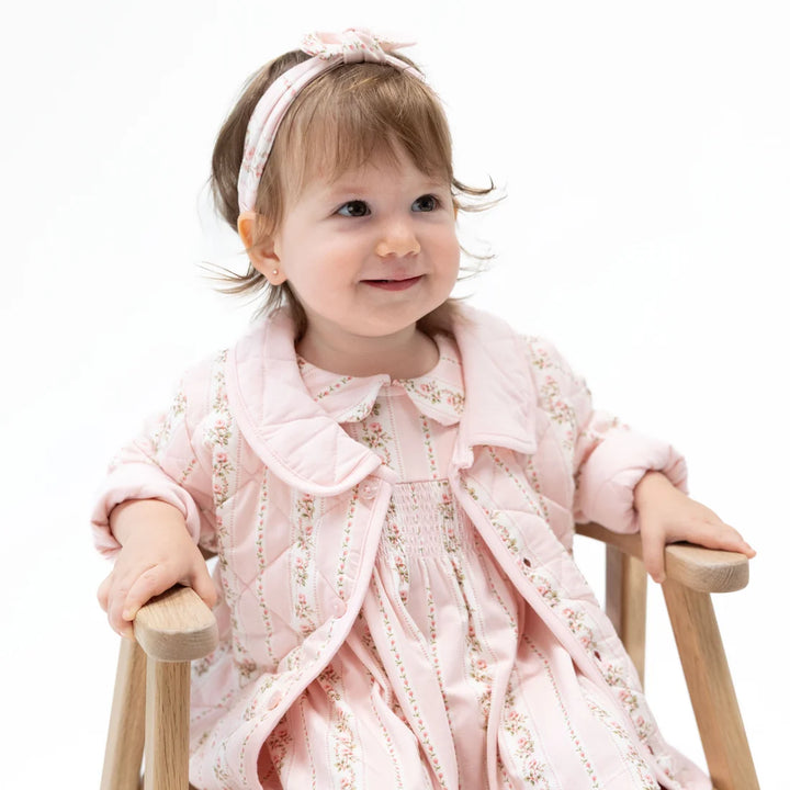 A baby girl wearing a light pink floral quilted jacket over a matching pink dress, sitting on a wooden chair against a white background.