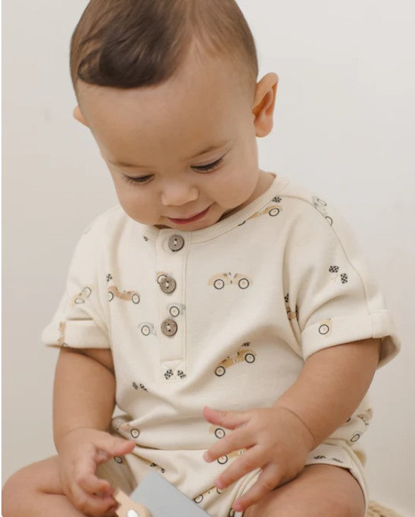 Baby wearing a cream-colored outfit with race cars and with buttons and small patterns, sitting on a light surface.