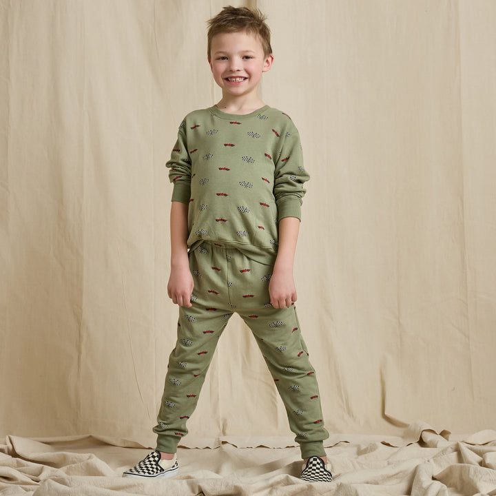 A young boy stands smiling in a matching sage green pajama set with a small repeating pattern, consisting of a long-sleeve top and jogger pants, against a beige fabric backdrop.