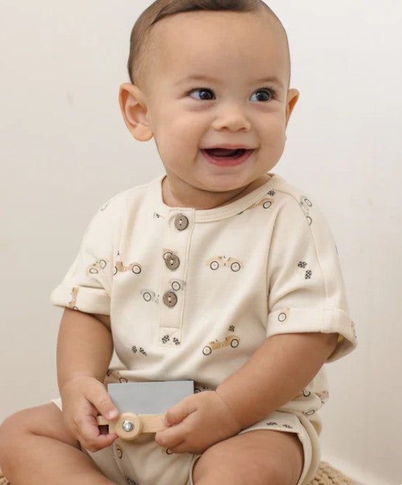 Baby wearing a cream-colored outfit with race car print with buttons on a plain background