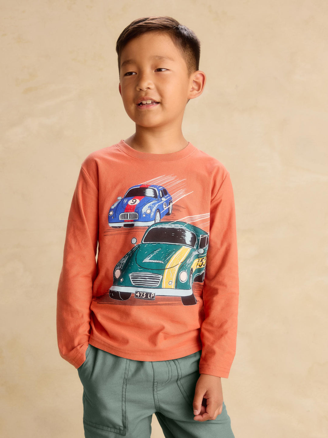 A young boy wearing an orange long-sleeve t-shirt with a graphic of two racing cars, paired with sage green pants, standing against a textured beige background.