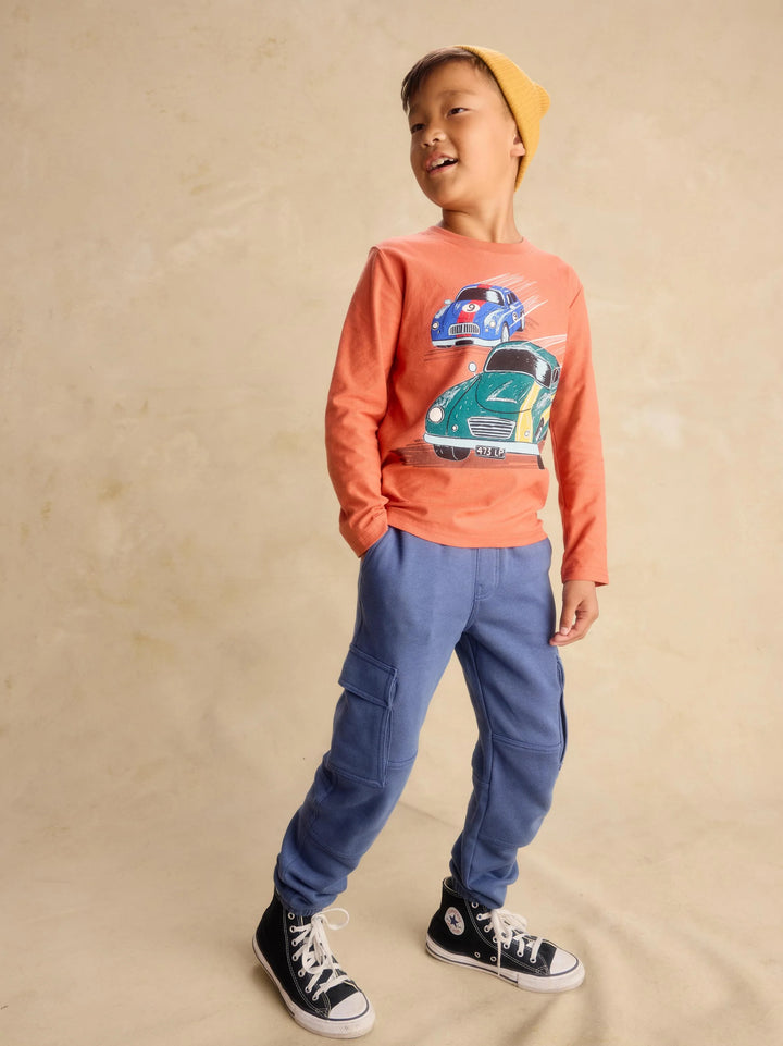 A young boy wearing an orange long-sleeve graphic t-shirt with a car print, blue cargo pants, black and white sneakers, and a yellow beanie, standing against a textured beige backdrop.