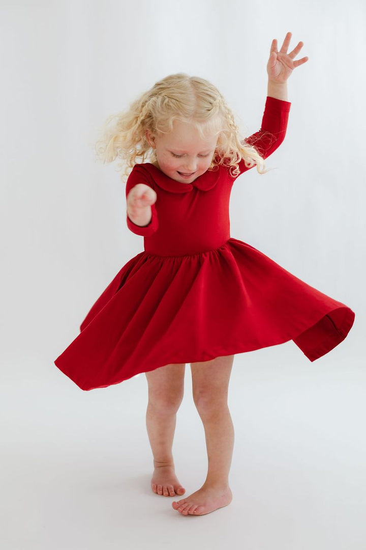 A young blonde girl with curly hair wearing a bright red long-sleeve dress with a Peter Pan collar and a flared skirt, captured mid-spin on a seamless white studio background.