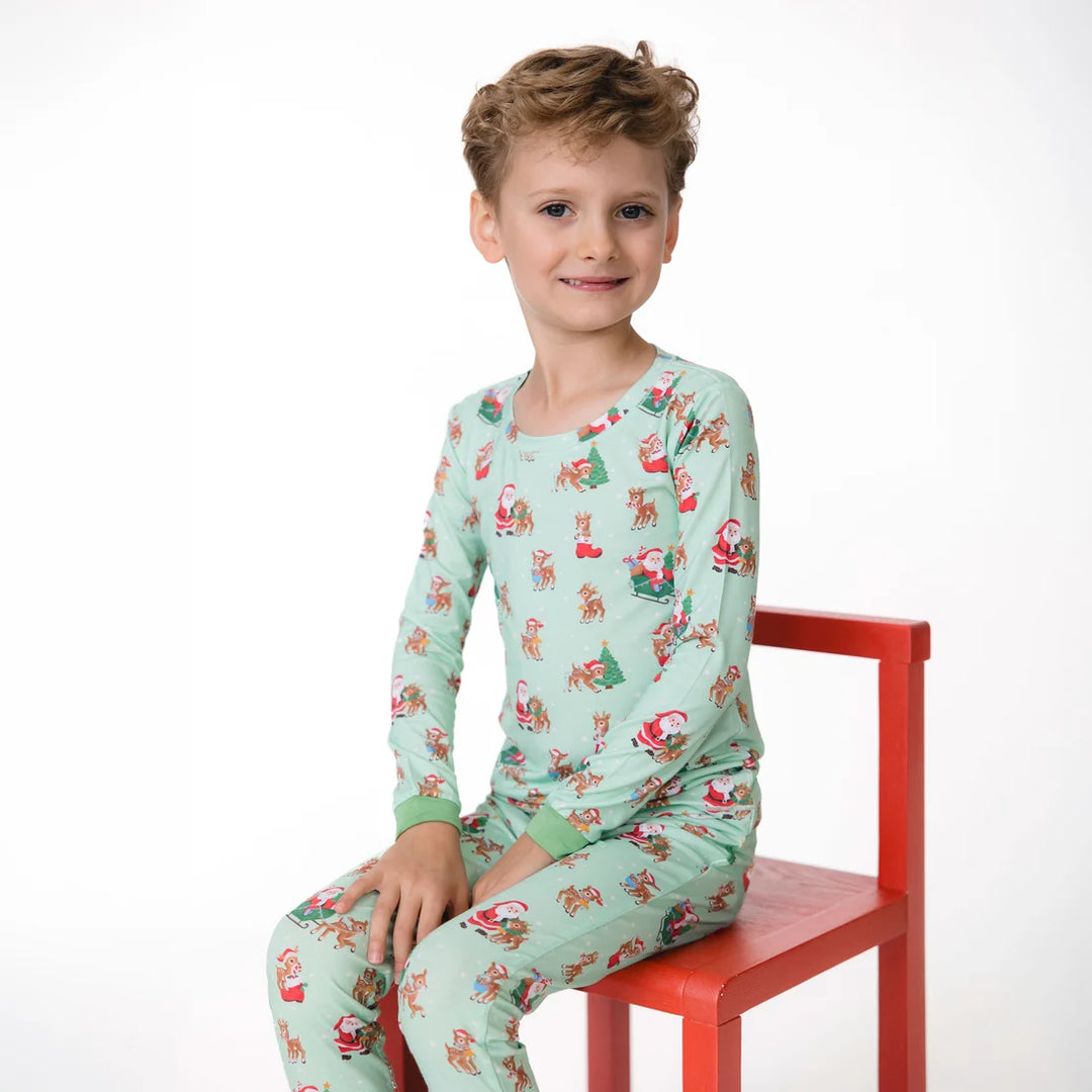 A young boy wearing a light mint green long-sleeve pajama set with a festive Santa Claus and Christmas tree print, sitting on a red wooden chair against a white studio background.