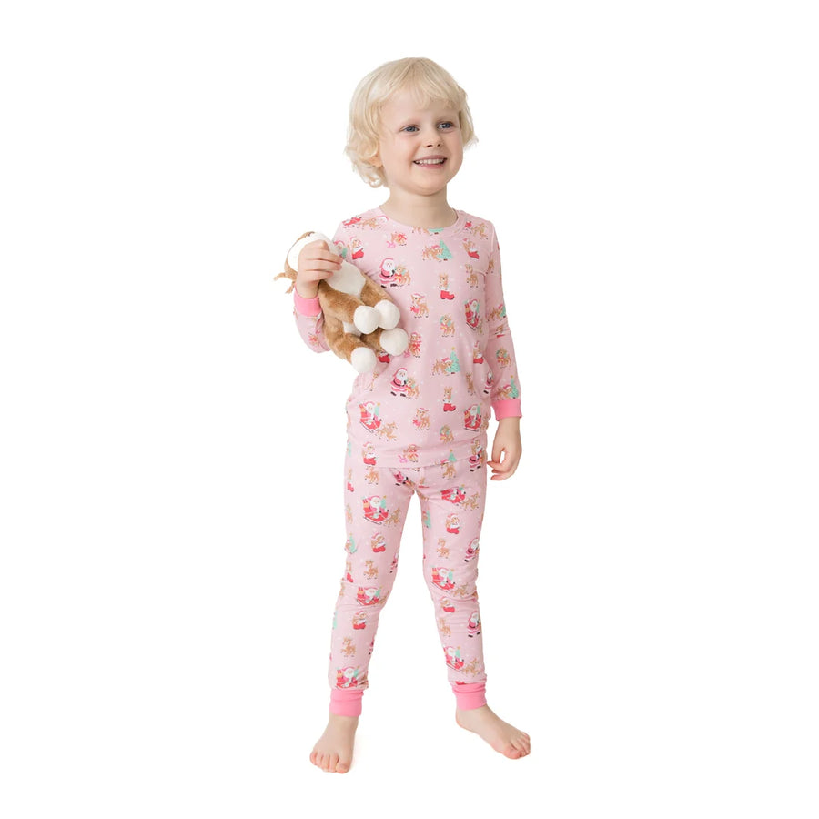 A young child with blonde hair stands barefoot against a white background, wearing a light pink long-sleeve pajama set with a colorful cartoon holiday pattern and holding a plush toy.