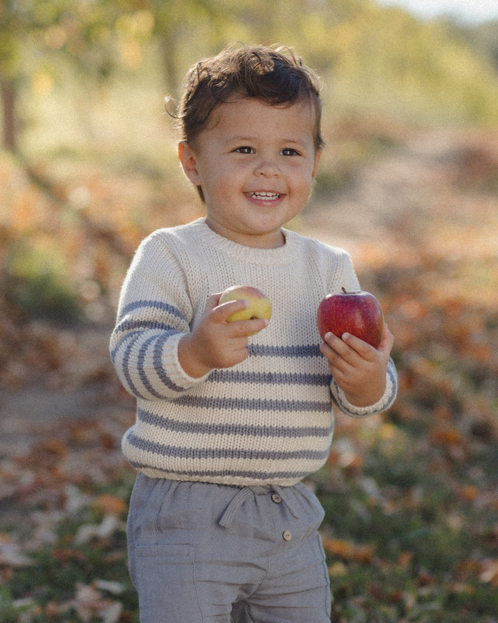 A smiling toddler standing outdoors in a blurred garden or orchard setting, wearing a cream knit sweater with gray horizontal stripes and light gray pants, holding a yellow apple in one hand and a red apple in the other.