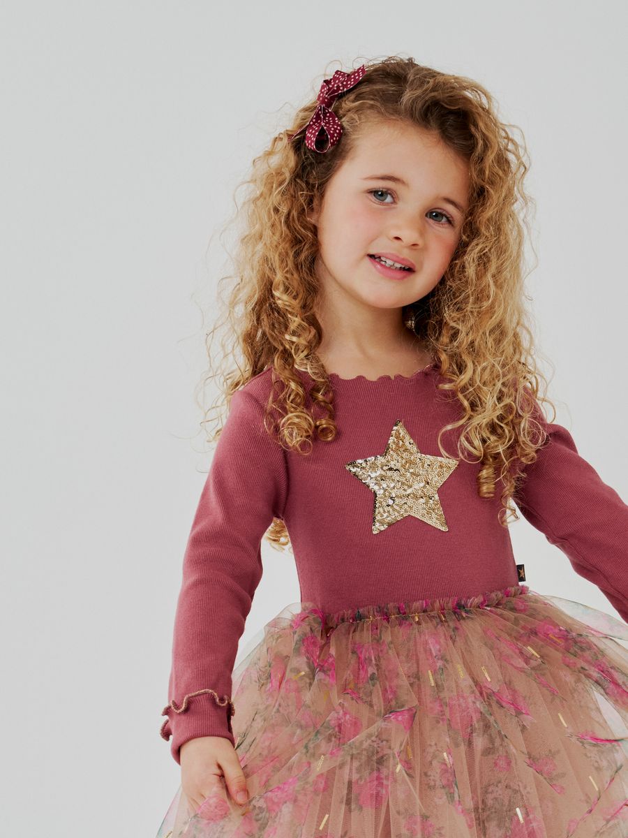 A young girl with curly blonde hair wearing a dusty rose long-sleeve top with a gold sequined star on the chest and a pink floral tulle tutu skirt, photographed against a light gray studio background.