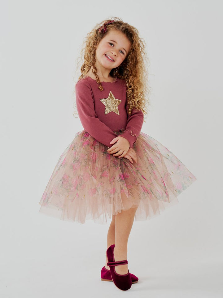 A young girl with curly light brown hair is modeling a two-piece outfit consisting of a dusty rose long-sleeve top with a gold sequin star and a light pink floral tulle tutu skirt. She is wearing burgundy velvet Mary Jane shoes against a plain light gray studio background.