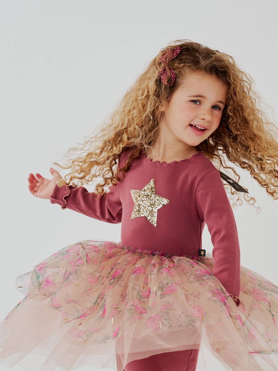 A young girl with long curly blonde hair wearing a dusty rose long-sleeve top with a glittery gold star on the chest and a pink floral tulle tutu skirt posed against a light gray studio background