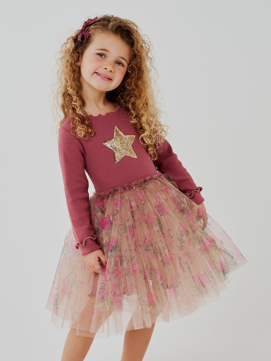 A young girl with curly light brown hair wearing a dusty rose long-sleeve top with a gold sequined star and a pink floral tulle tutu skirt, standing against a light gray studio background.