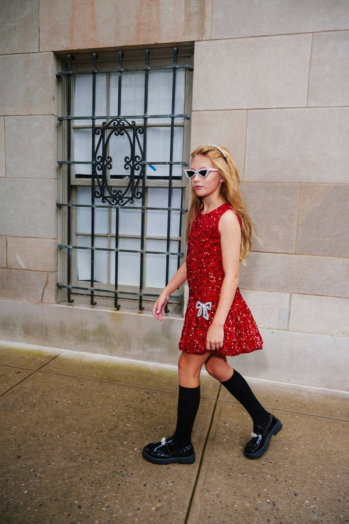 A young girl with blonde hair walks along a sidewalk wearing a sleeveless red sequined dress with a white bow, black knee-high socks, and black shoes, standing in front of a stone wall with a barred window.