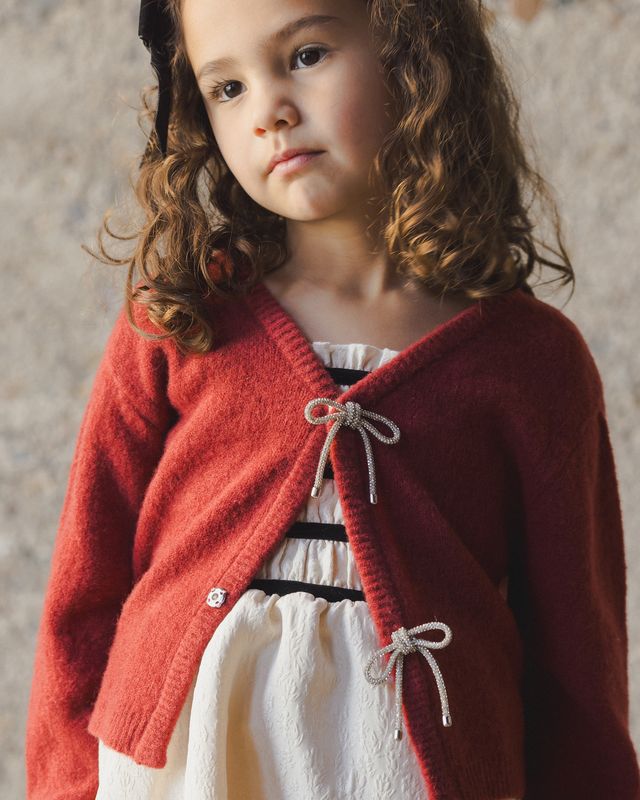 Young girl wearing a red knitted cardigan over a white dress against a neutral background