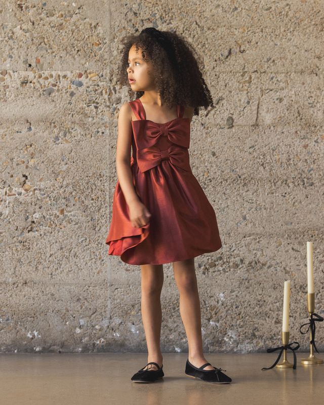 Young girl in a red dress standing against a textured wall with candles on a stand.