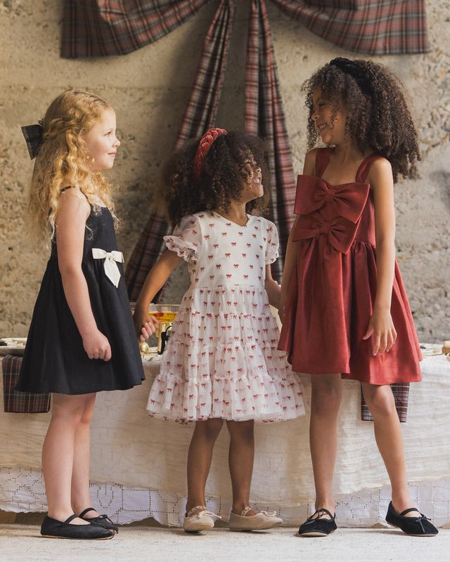 Three young girls in dresses standing together in a room with plaid curtains.