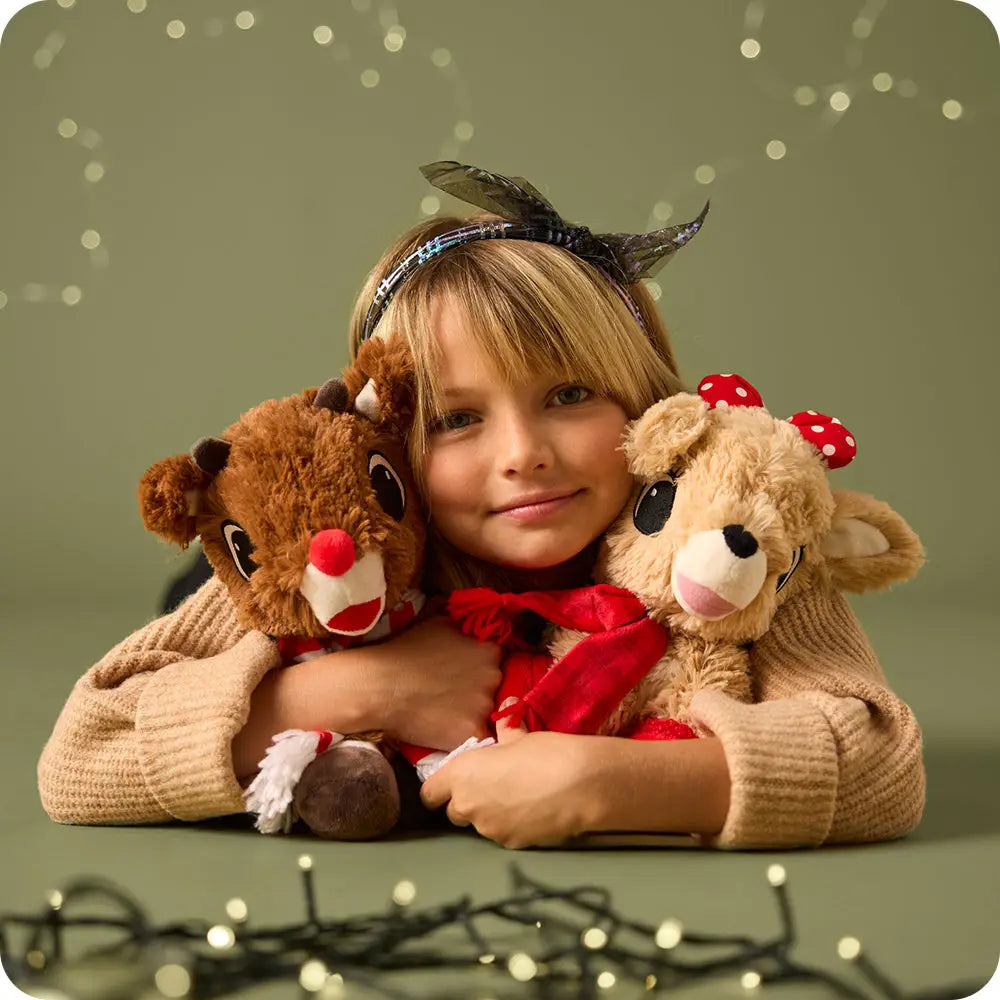A young girl with blonde hair wearing a reindeer antler headband hugs two plush teddy bears, one brown and one beige, against a soft olive green background with decorative string lights.