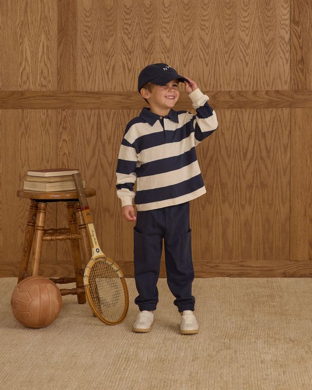 Child wearing a striped shirt and cap, standing next to a stool with books and sports equipment against a wooden paneled wall.