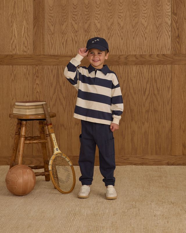Child wearing a striped shirt and cap, standing next to a wooden stool and vintage tennis racket against a wooden paneled wall.