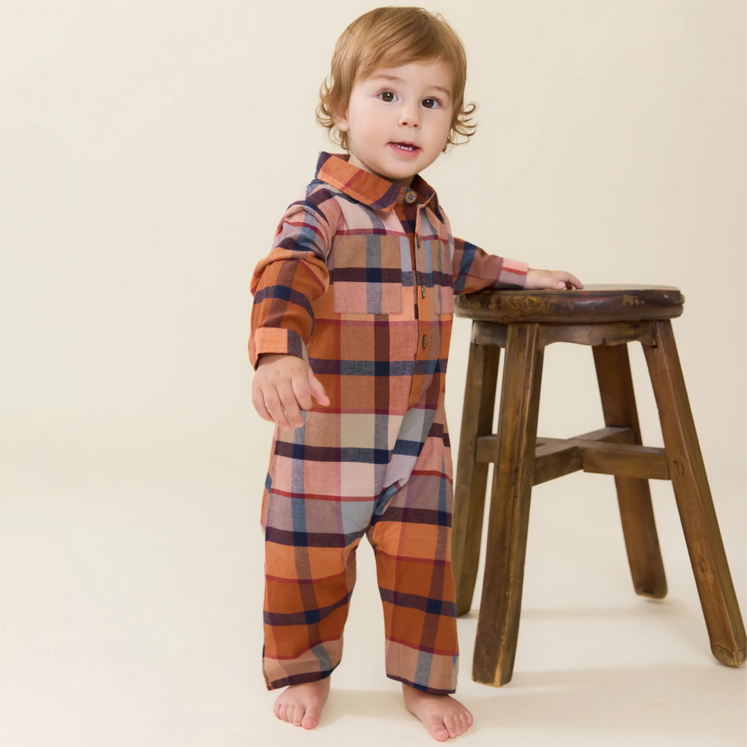 baby boy wearing a rust plaid jumpsuit standing next to a stool. 