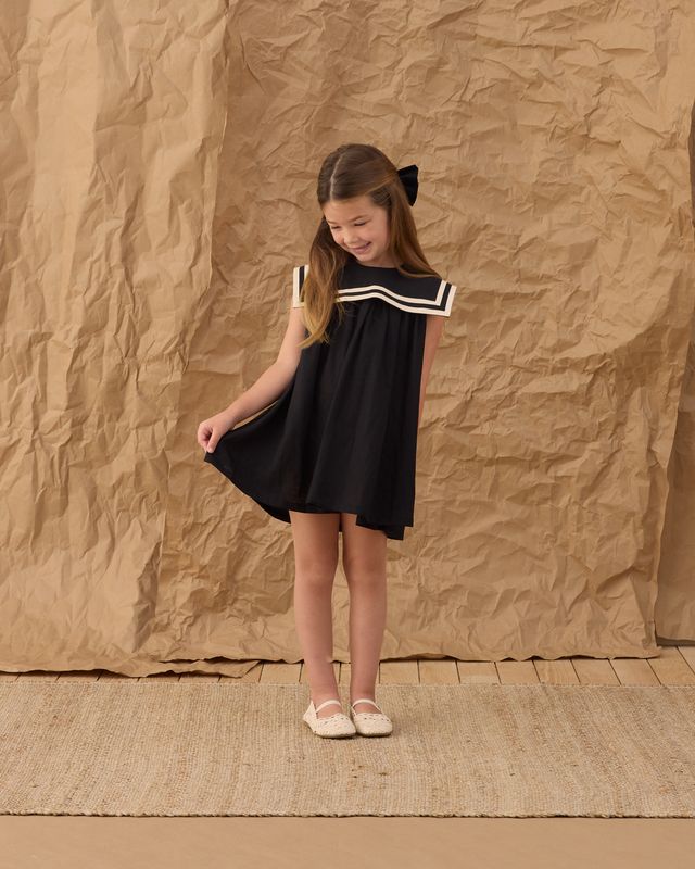 young girl pulling slightly at her skirt in a black sailor dress with white piping. 