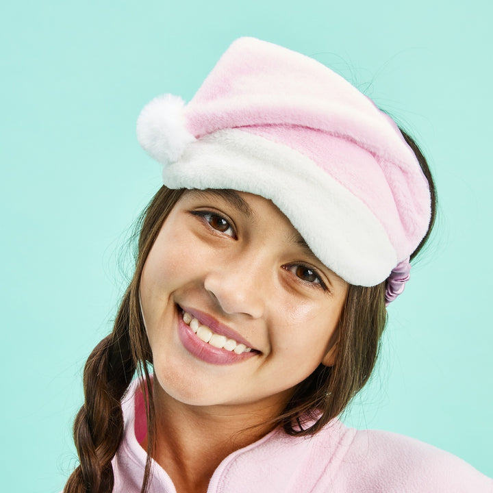 A smiling young girl wearing a plush pink and white Santa hat with a fluffy pom-pom, posed against a solid light aqua background.
